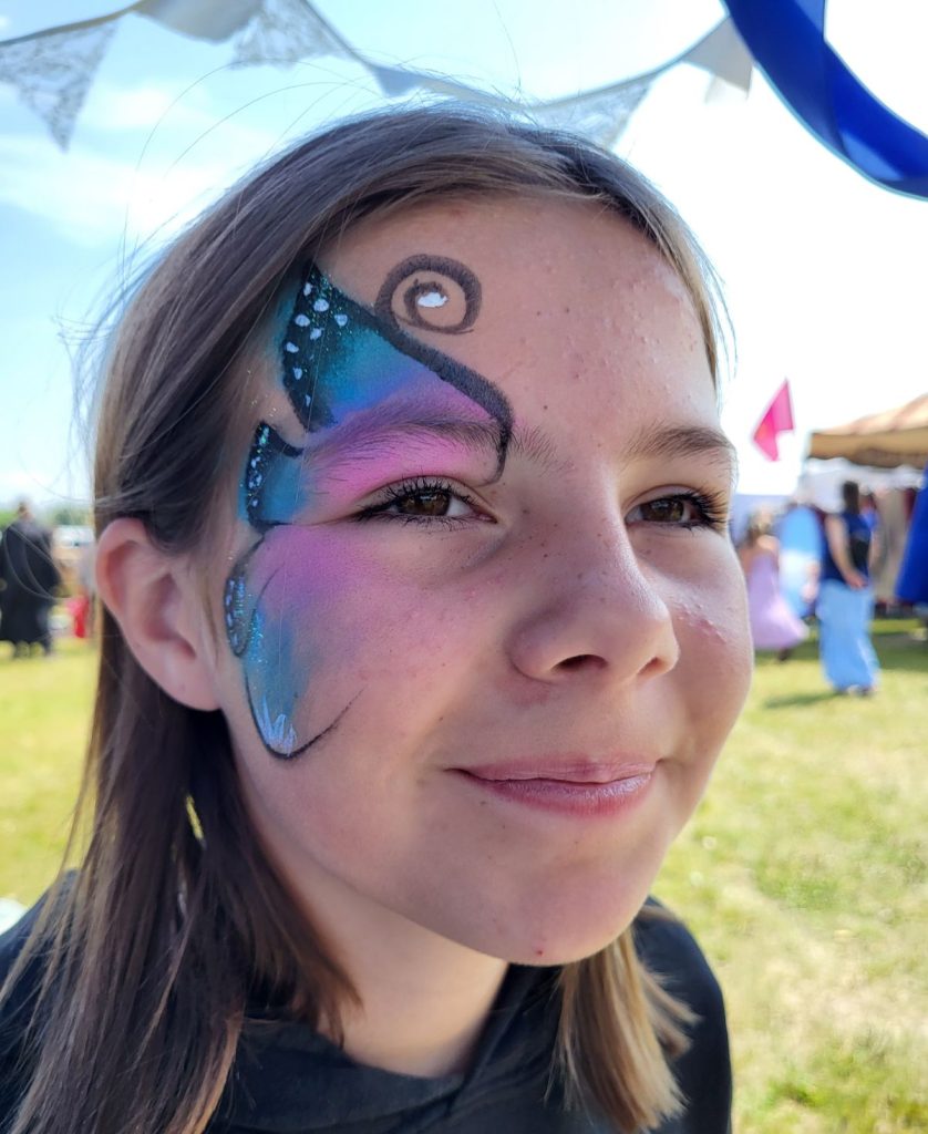 A smiling girl looking off to the right of the frame with a big butterfly wing on the closest side of her face that is blue, purple and black.